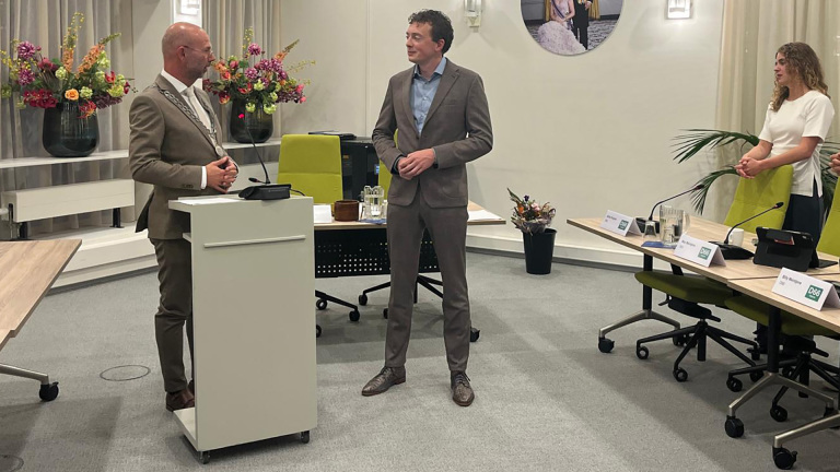 Man met ambtsketen spreekt bij lessenaar in vergaderzaal, terwijl man in pak en vrouw toekijken