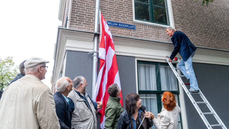 Man op ladder hangt straatnaambord Willem Rederplaats op, terwijl omstanders toekijken bij hoekpand met vlag