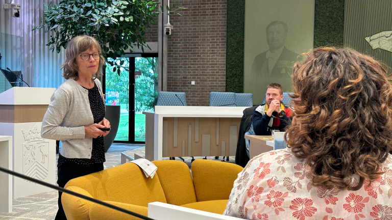 Vrouw spreekt in een raadszaal, met een man aan tafel en een toehoorder op de voorgrond