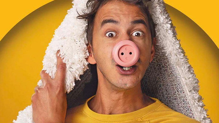 Man met verbaasde blik en roze varkensneus, met een pluizige deken over zijn hoofd tegen gele achtergrond