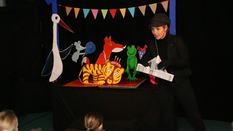 Verteller bij poppentheater met gekleurde dierfiguren (ooievaar, muis, vos, kikker, tijger) onder slingers