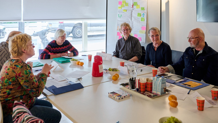 Groep ouderen in overleg rond een tafel met papieren, bekers en fruit, in een lichte vergaderruimte met post-its op de muur