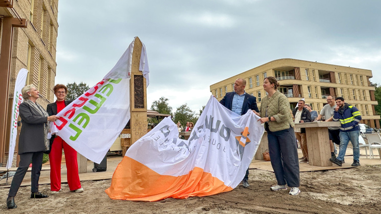 Mensen onthullen met witte doeken een houten insectenhotel op een plein, met toeschouwers en gebouwen op de achtergrond