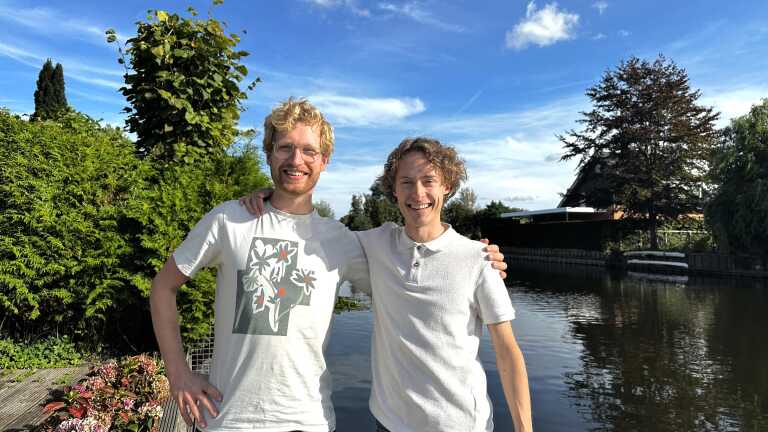 Twee lachende mannen met hun armen om elkaar, staand bij een vijver met bomen en blauwe lucht op de achtergrond