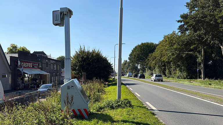 Flitspaal langs een provinciale weg met passerende auto’s, naast groen bermgebied en gebouwen met het bord Casper