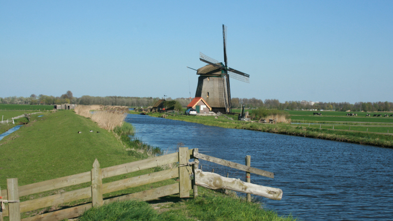 Een Nederlandse polderlandschap met een tradionele molen naast een kanaal, omgeven door groene velden en bomen.