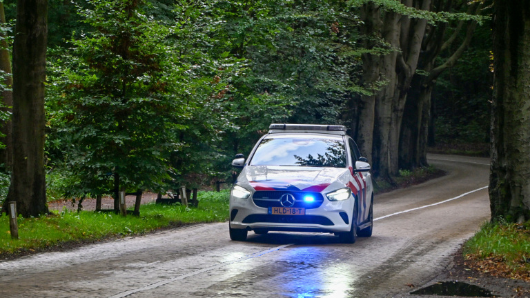 Nederlandse politieauto met zwaailicht rijdt over een natte weg door een bosrijk gebied
