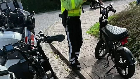 Persoon in fluoriserend hesje staat op stoep tussen een motor en een bromfiets langs de straat