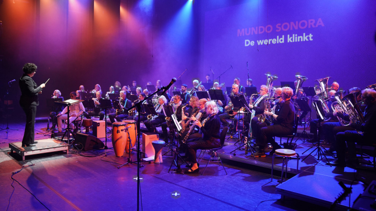 Dirigent leidt een groot blaasorkest op podium met paarsblauwe belichting; op achterwand staat “MUNDO SONORA De wereld klinkt”