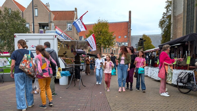 Drukke straatmarkt met kramen en winkelend publiek, met een viswagen en vlaggen met tekst Hoorn en Hollands
