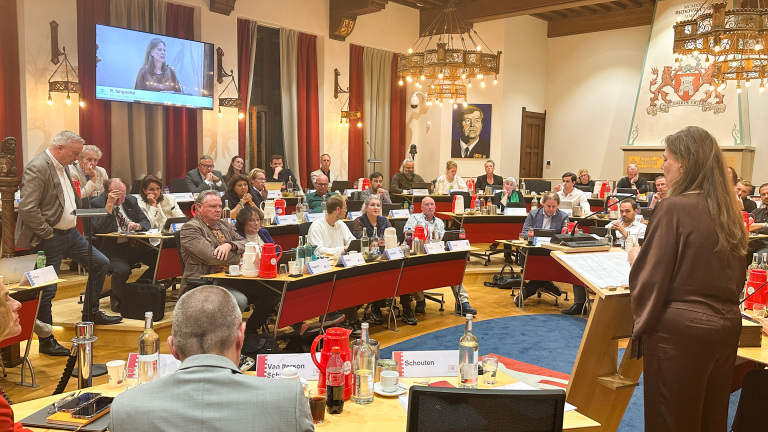 Raadsvergadering in een grote zaal; vrouw spreekt vanaf katheder terwijl raadsleden aan tafels luisteren en noteren