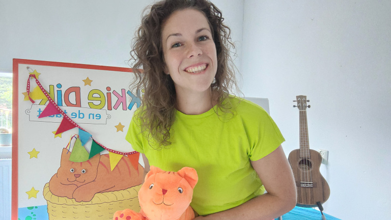Lachende vrouw in felgroen shirt houdt een oranje knuffelkat vast, met kinderposter en ukelele op de achtergrond