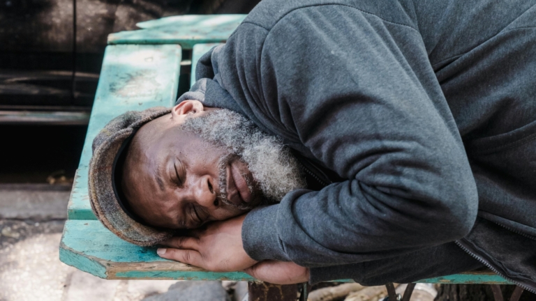 Man sleeping on a bench in a city, next to a cardboard sign reading 