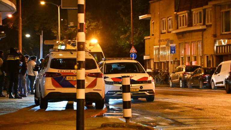Twee politieauto’s met zwaailichten op een natte straat in de avond, met omstanders op de stoep