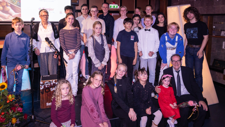 Groepsfoto van kinderen en volwassenen op een podium, met microfoons, bloemen en een projectiescherm op de achtergrond