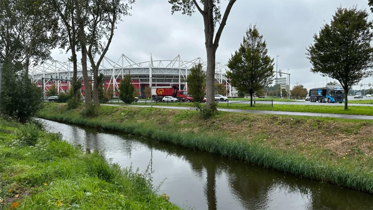 Stadion achter bomen langs een weg, met vrachtwagens en auto’s naast een sloot in het groen