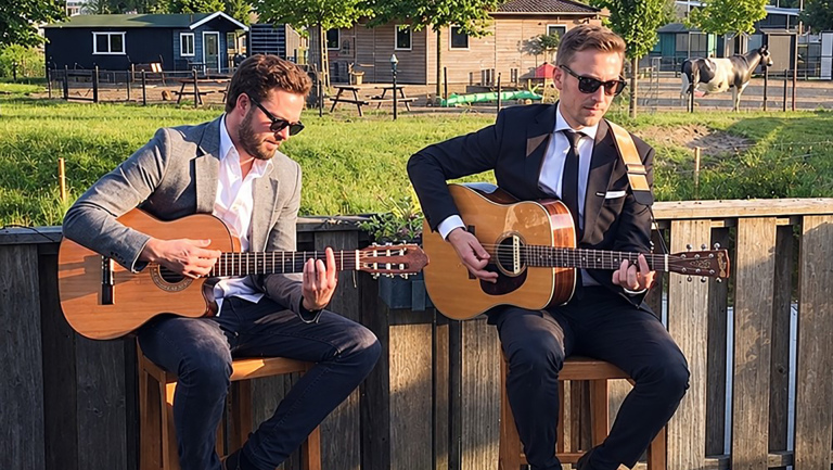 Twee mannen met zonnebril spelen akoestische gitaar op krukken buiten, met een boerderij en paard op de achtergrond
