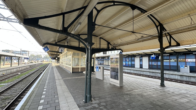 Leeg perron 6 op een treinstation met overkapping, spoorrails, prullenbakken en een digitaal informatiebord