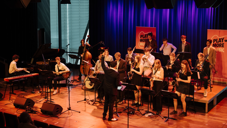 Bigband op podium met dirigent, piano, gitaar, contrabas, drums en blazers voor banners met tekst Play More Music