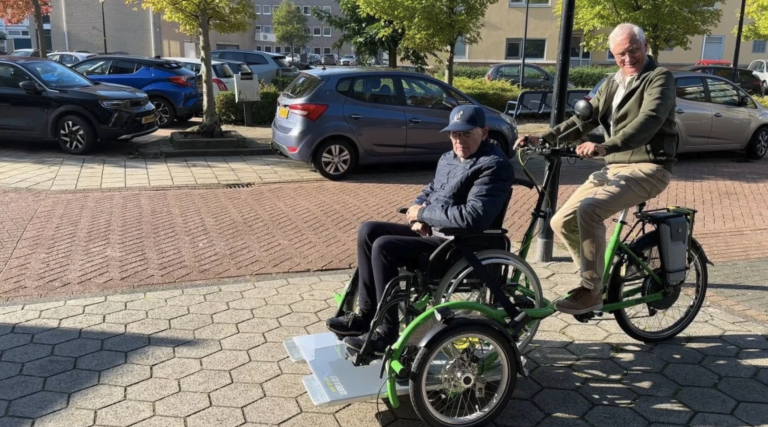Man op groene duofiets met rolstoelplatform, met passagier in rolstoel, op straat bij geparkeerde auto’s