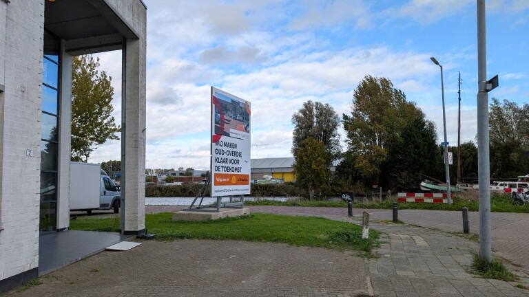 Informatiebord langs het water met tekst “Wij maken Oud-Overdie klaar voor de toekomst” naast een gebouw en weg