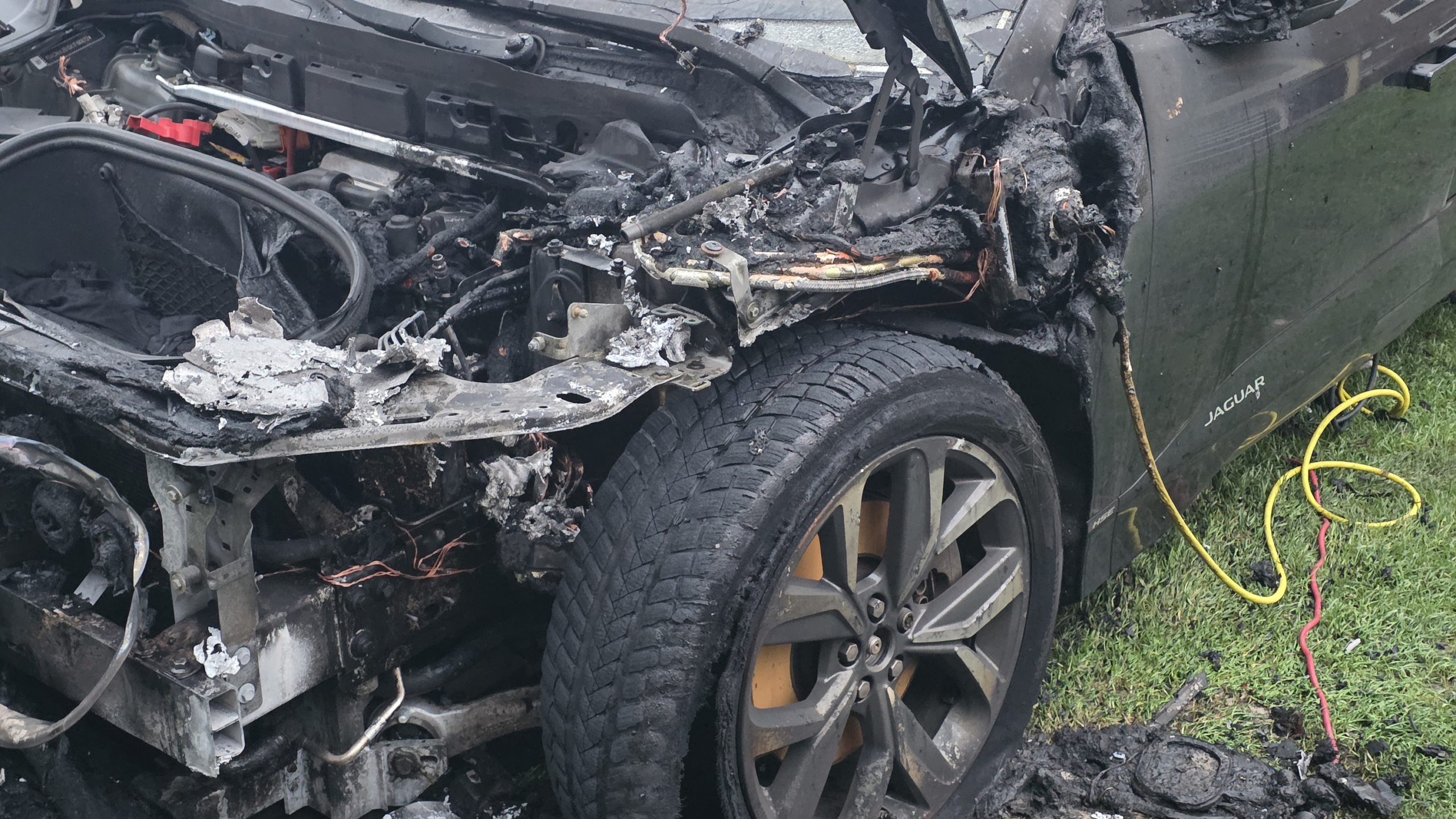 Auto met uitgebrand motorcompartiment en open motorkap op een grasveld, omringd door gebouwen en een geparkeerde witte auto.