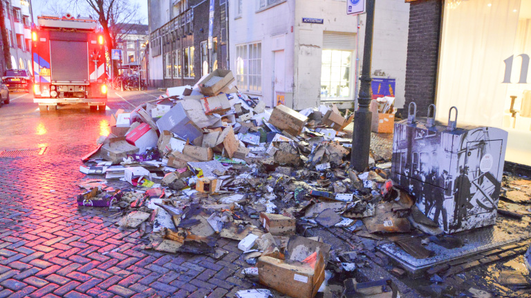 Afvalhopen van kartonnen dozen en puin op een natte straat naast een brandweerwagen en dicht bij een winkel.