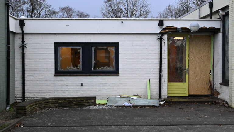 Vervallen gebouw met gebroken ramen en een dichtgetimmerde deur, omgeven door kapotte materialen op de grond.