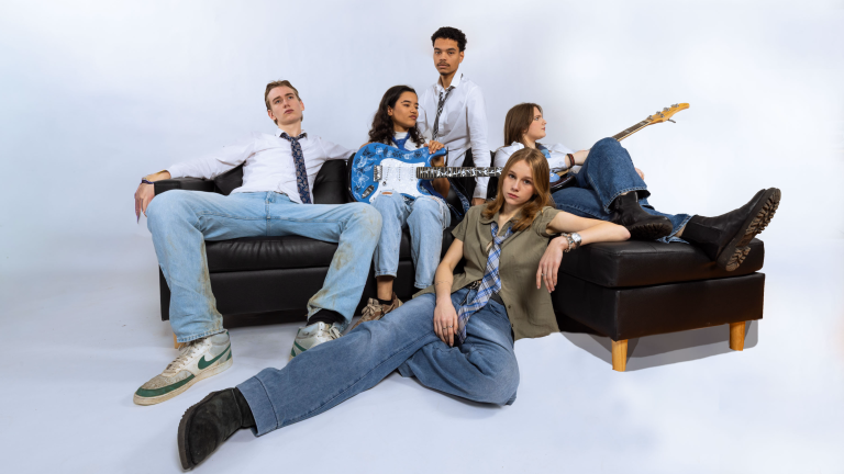 Vijf mensen zitten nonchalant op twee zwarte banken, sommigen met gitaren, gekleed in casual outfits.