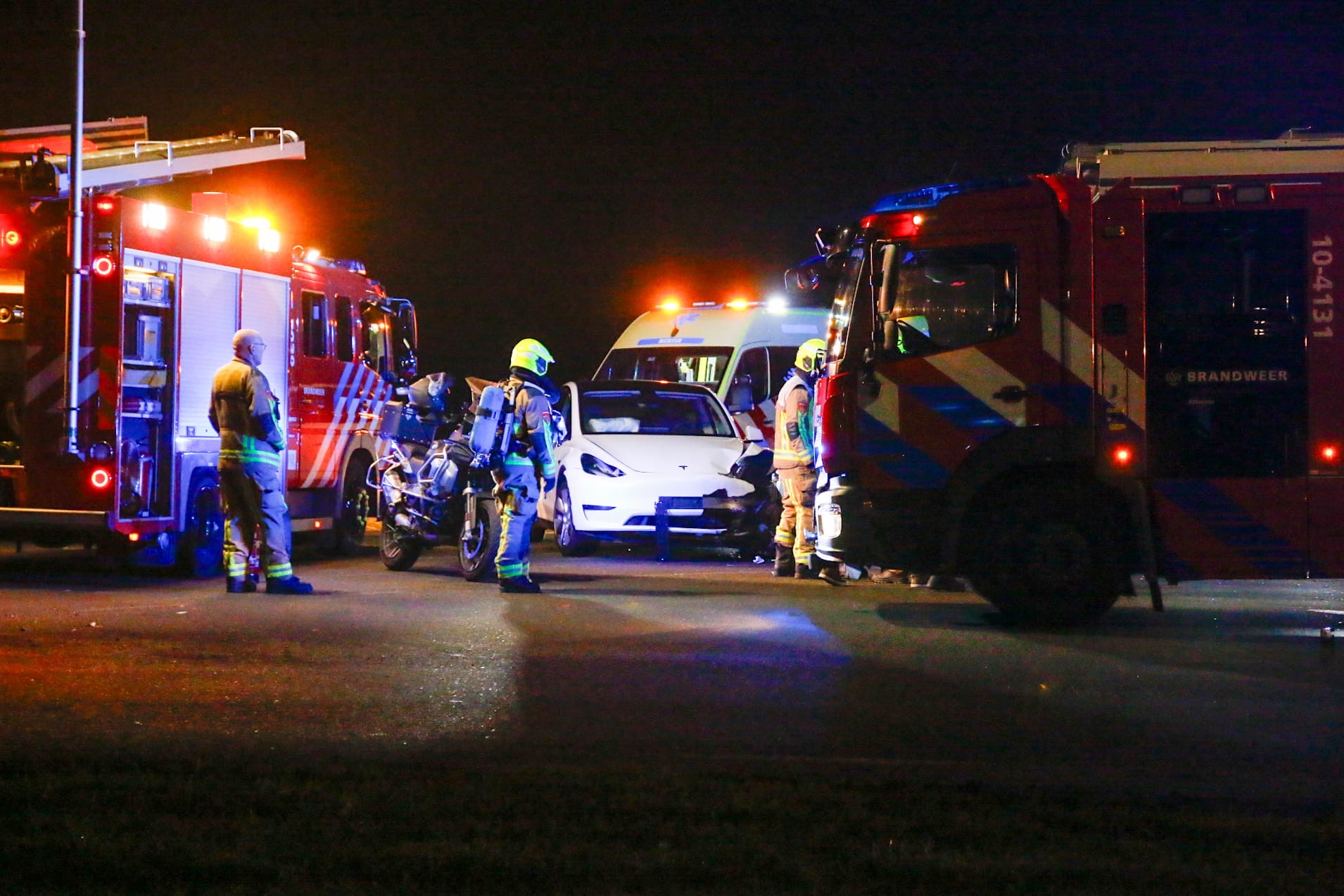 Een blauw beschadigde auto staat langs de weg bij nacht, omringd door hulpverleners en voertuigen met knipperende lichten.