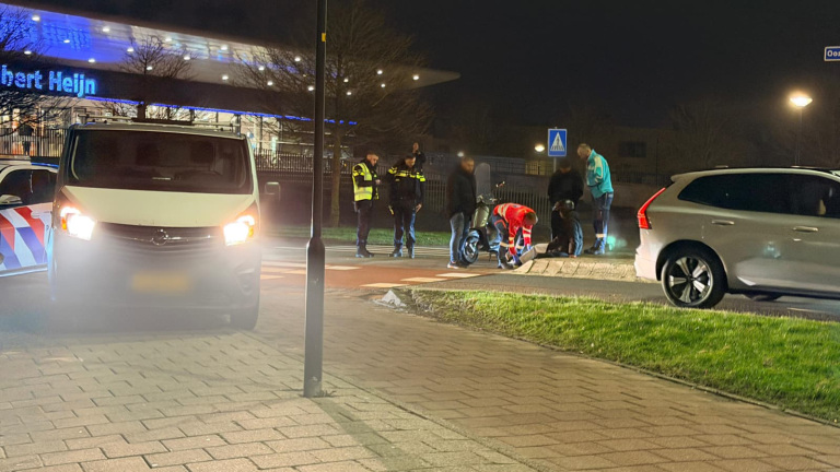 Ambulancepersoneel verzorgt iemand op een brancard naast een scooter bij een rotonde, met een ambulance op de achtergrond.