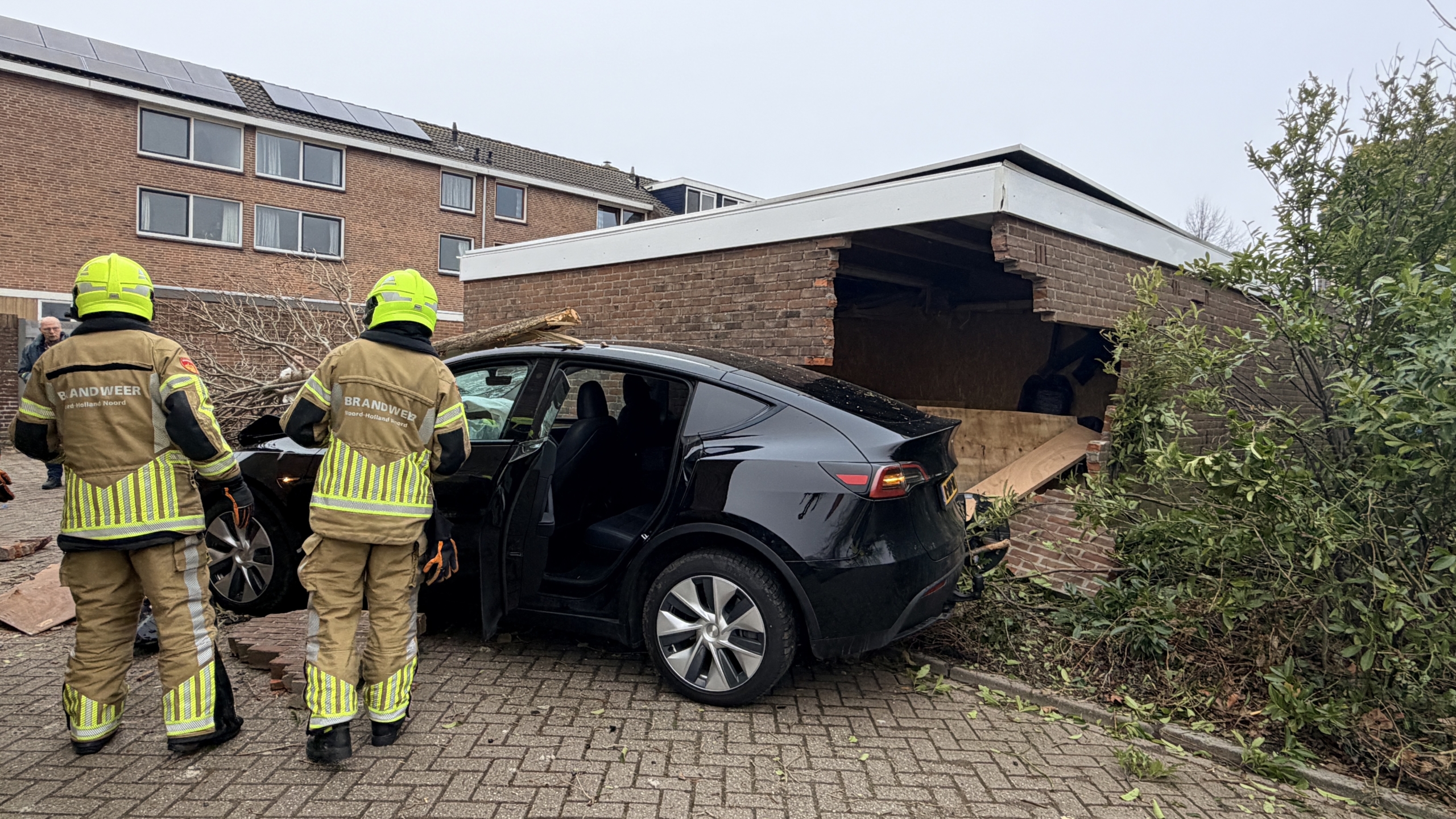 Een auto is gecrasht in een garage, omringd door puin en een omgevallen boom, met een gele reddingswagen op de achtergrond.