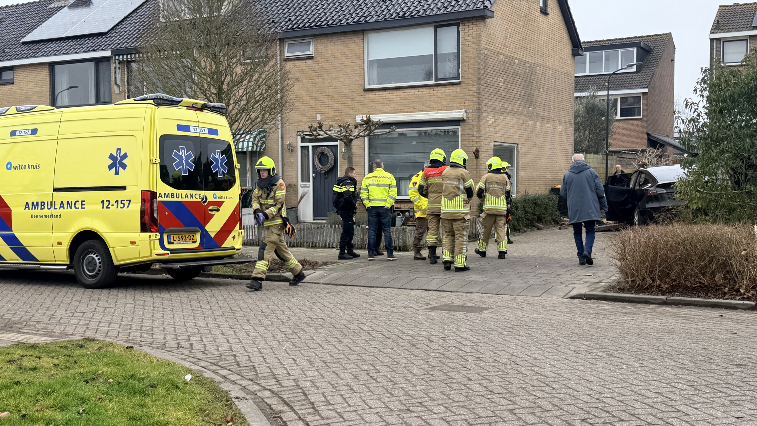 Een auto is gecrasht in een garage, omringd door puin en een omgevallen boom, met een gele reddingswagen op de achtergrond.