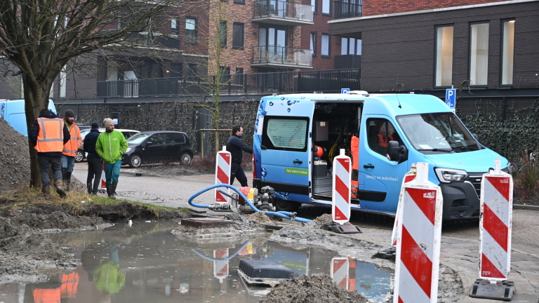 Bouwvakkers werken naast een blauw servicevoertuig rond een modderige waterplas, omgeven door waarschuwingsborden.