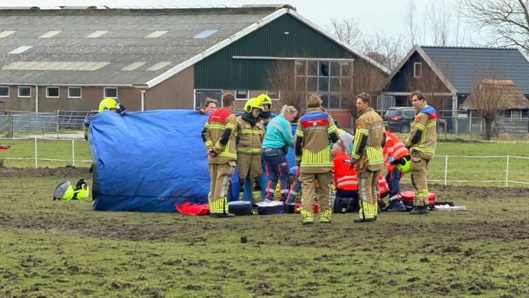 Hulpdiensten, waaronder brandweerlieden en medisch personeel, werken samen in een veld bij een boerderij.