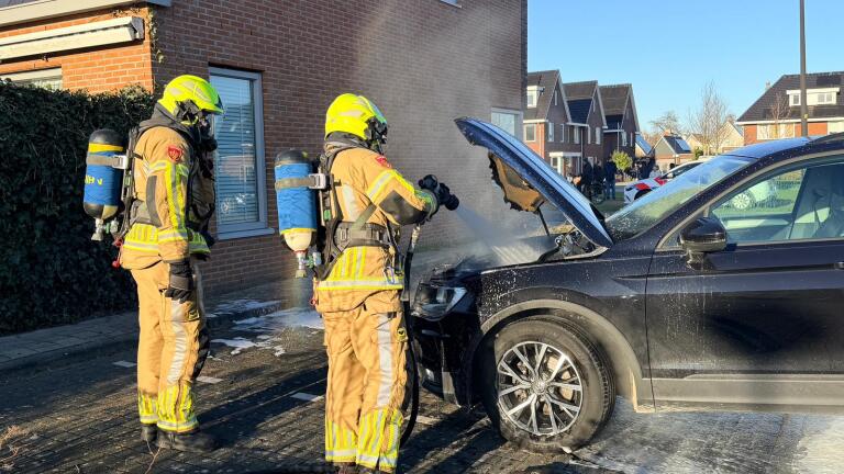 Brandweerlieden blussen een brand in de motorkap van een zwarte auto in een woonwijk.