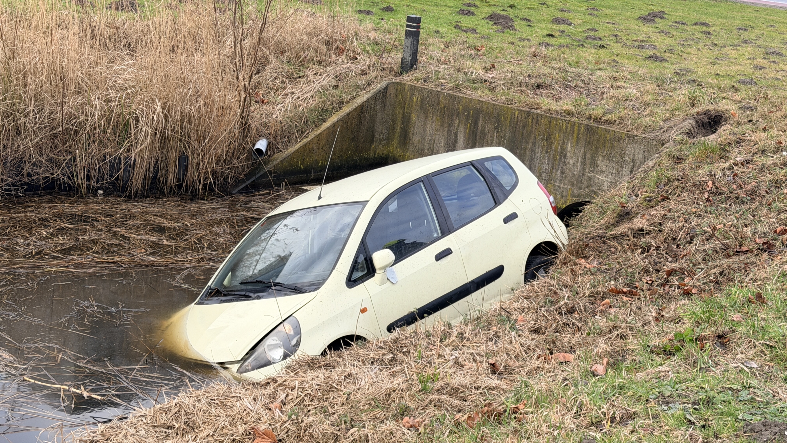 Auto half ondergedompeld in een sloot, met brandweer, ambulances en mensen op de achtergrond.