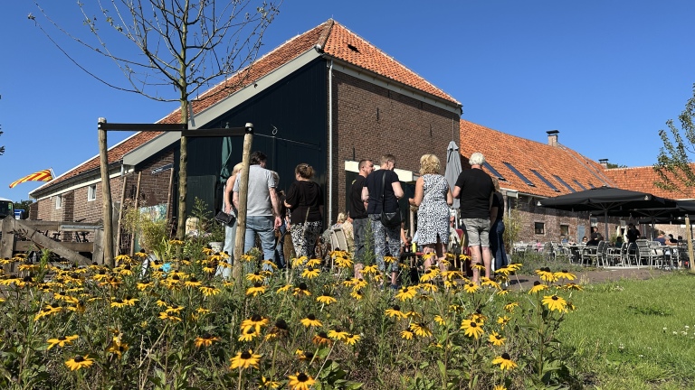 Groep mensen staat buiten bij een gebouw met een rood dak, omringd door bloeiende gele bloemen onder een heldere blauwe lucht.