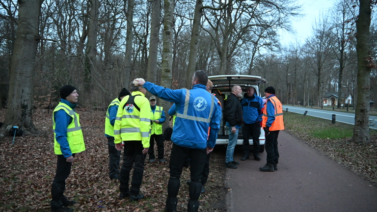 Mensen in gele veiligheidsvesten lopen door een bos met herfstbladeren op de grond, naast een busje.