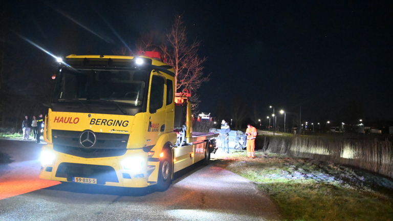 Auto ligt op zijn zij in het gras langs een weg, omgeven door straatlantaarns en een gebouw op de achtergrond, 's nachts.