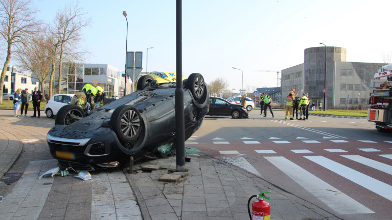 Auto ligt ondersteboven op straat na ongeval, omringd door hulpverleners, met een brandblusser op de voorgrond.