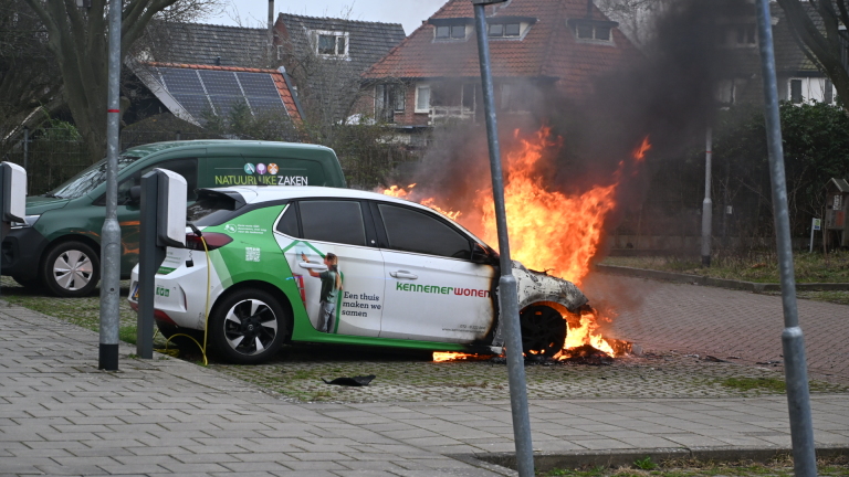 Brandweerlieden blussen een elektrische auto die in brand staat bij een laadpaal, omgeven door rook.