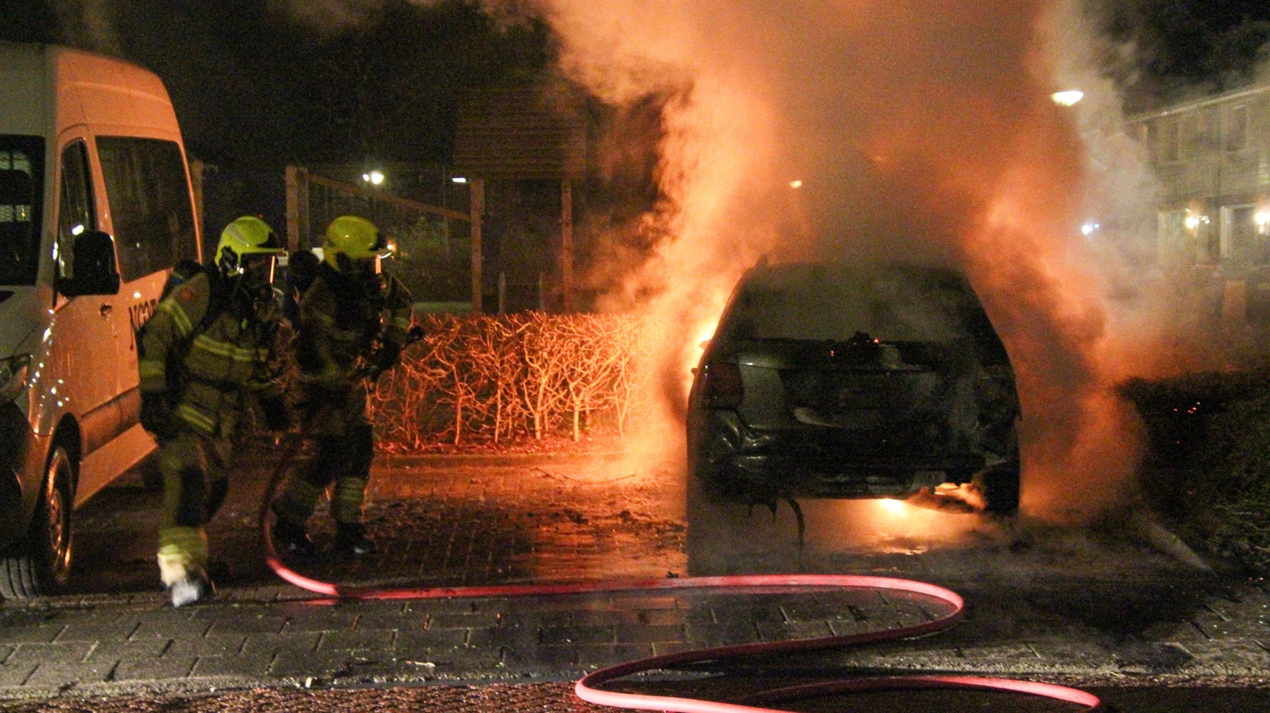 Auto in brand met grote vlammen, naast een boom en een andere geparkeerde auto in een nachtelijke omgeving.