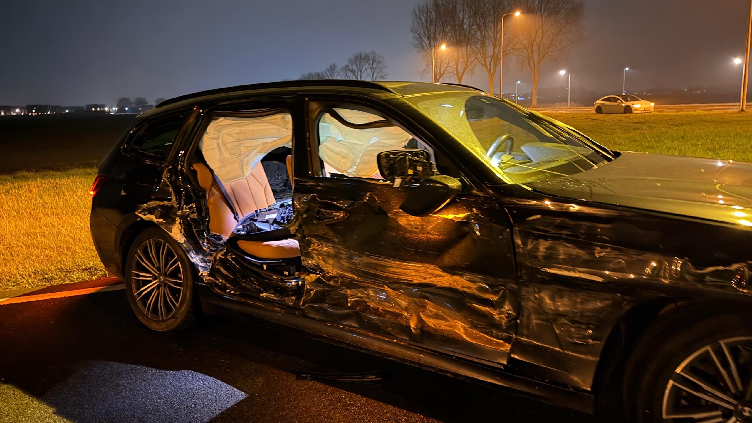 Auto met deukschade en geopende airbags bij een rotonde 's nachts, met verzamelde mensen en kijkers op de achtergrond.