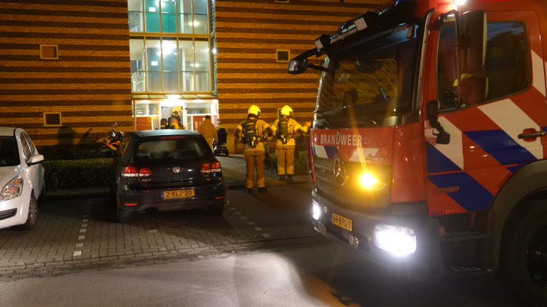 Brandweerlieden in actie bij een appartementengebouw met een brandweerwagen op de voorgrond en auto's op de parkeerplaats.