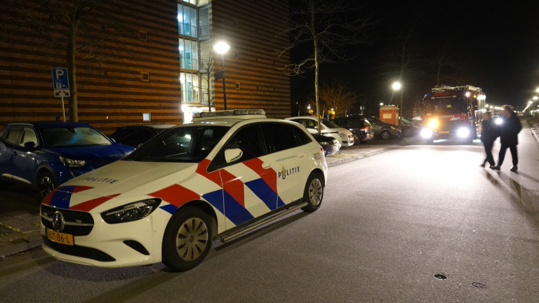 Brandweerlieden in actie bij een appartementengebouw met een brandweerwagen op de voorgrond en auto's op de parkeerplaats.