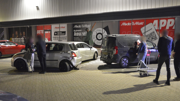 Twee auto's met schade op een parkeerplaats voor een MediaMarkt filiaal, omringd door mensen.