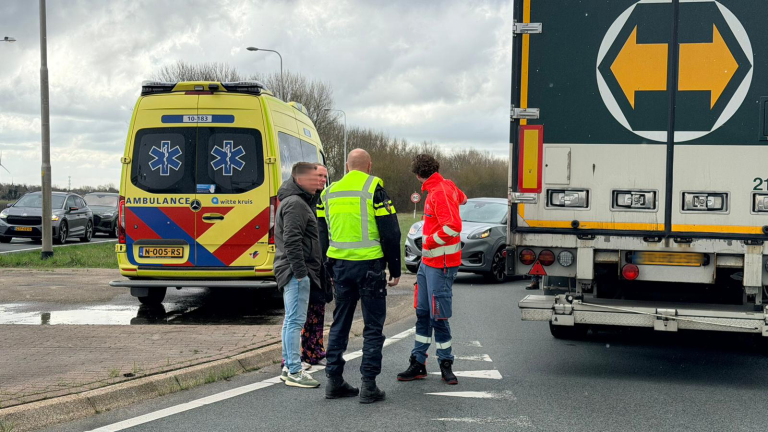 Een vrachtwagen, auto en ambulance staan stil bij een verkeerslicht. Mensen staan naast de voertuigen in gesprek.