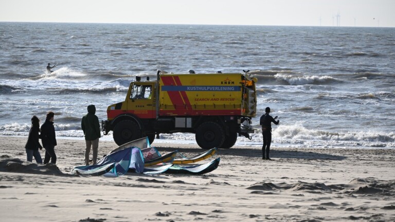 Kusthulpverleningsvoertuig van de KNRM op een strand met personen en een kitesurfer op zee op de achtergrond.