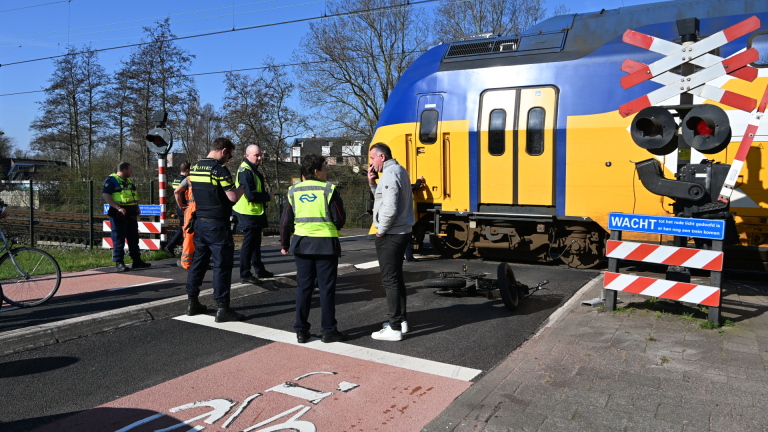 Een gele en blauwe passagierstrein staat bij een spoorwegovergang met politie- en spoormedewerkers op de voorgrond.